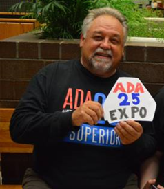 John Nousaine smiling and sitting on a bench at the ADA 25 Expo holding an Expo sign.