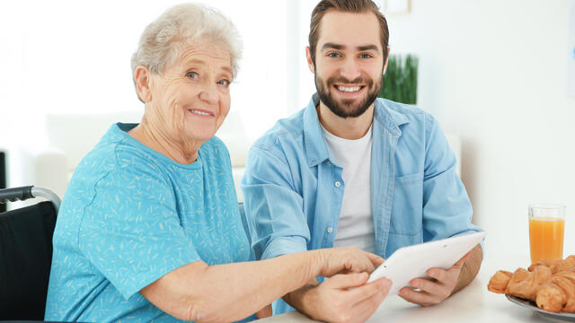 Caregiver holding a tablet sitting with an older adult at a table