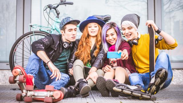 Four teens with skates, skateboard, bike taking a selfie while sitting on sidewalk