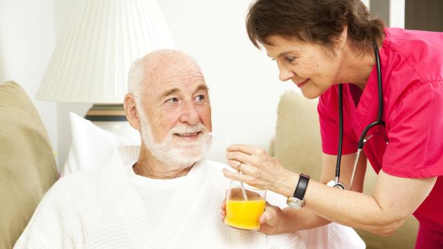 Elderly person in a nursing home is fed orange juice by a nurse