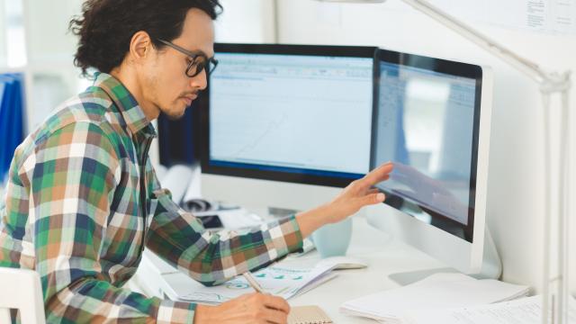 An adult making notes in a notebook at a desk with a computer monitor.