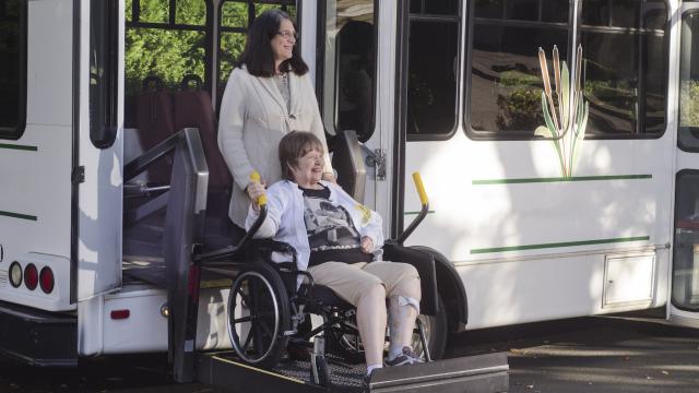 Person in a wheelchair being helped off the white bus lift by another person