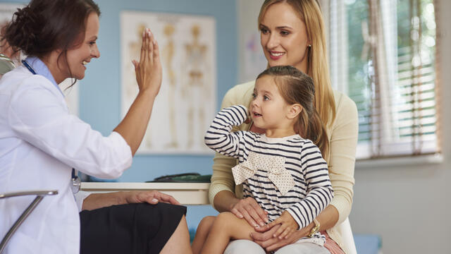 Doctor and young child sitting on an adult's lap exchange a high-five at a clinic