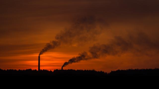 Exhaust cloud rises from a factory with red sky and sun.