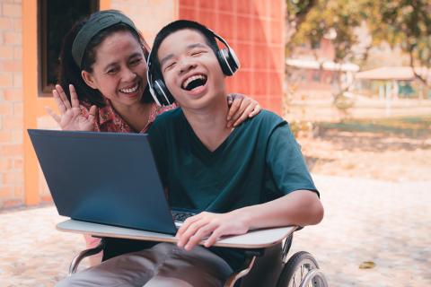 Child wearing headphones in a wheelchair with a laptop laughing with an adult