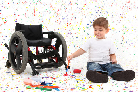 Child on the floor splatters paint a white room with a wheelchair close by