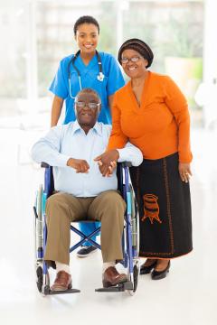 A man in a wheelchair holding hands with his wife and a healthcare provider standing behind, all smiling