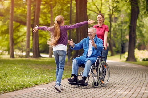 A teen greeting a grandparent in wheelchair being pushed by an adult outside.