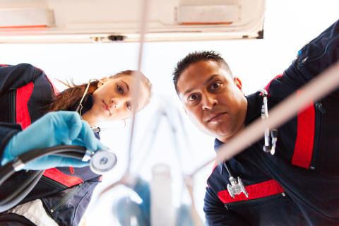 Two paramedics wearing blue suits with red stripes in ambulance, left side woman using stethoscope to provide first aid with a man on other side looking on and stethoscope around his neck.