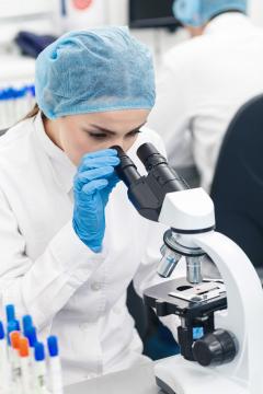 Scientist in blue hat, blue gloves and white lab coat examining through a microscope in a lab