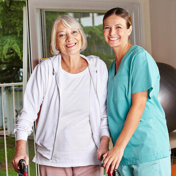 A smiling caregiver standing next to a smiling older adult with a walker.