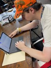 Man in an orange and green camouflage hat with a cream shirt sitting at a table using a computer