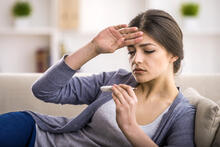 Young person on sofa, reading a thermometer while checking her forehead