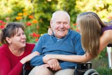 Young person hug elder person in a wheelchair with a person kneeling on other side of the wheelchair outside.