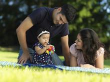 Two adults watching a baby playing with baby toys outside.