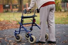 close up view of wheeled walker with person in tan top and pants behind on sidewalk